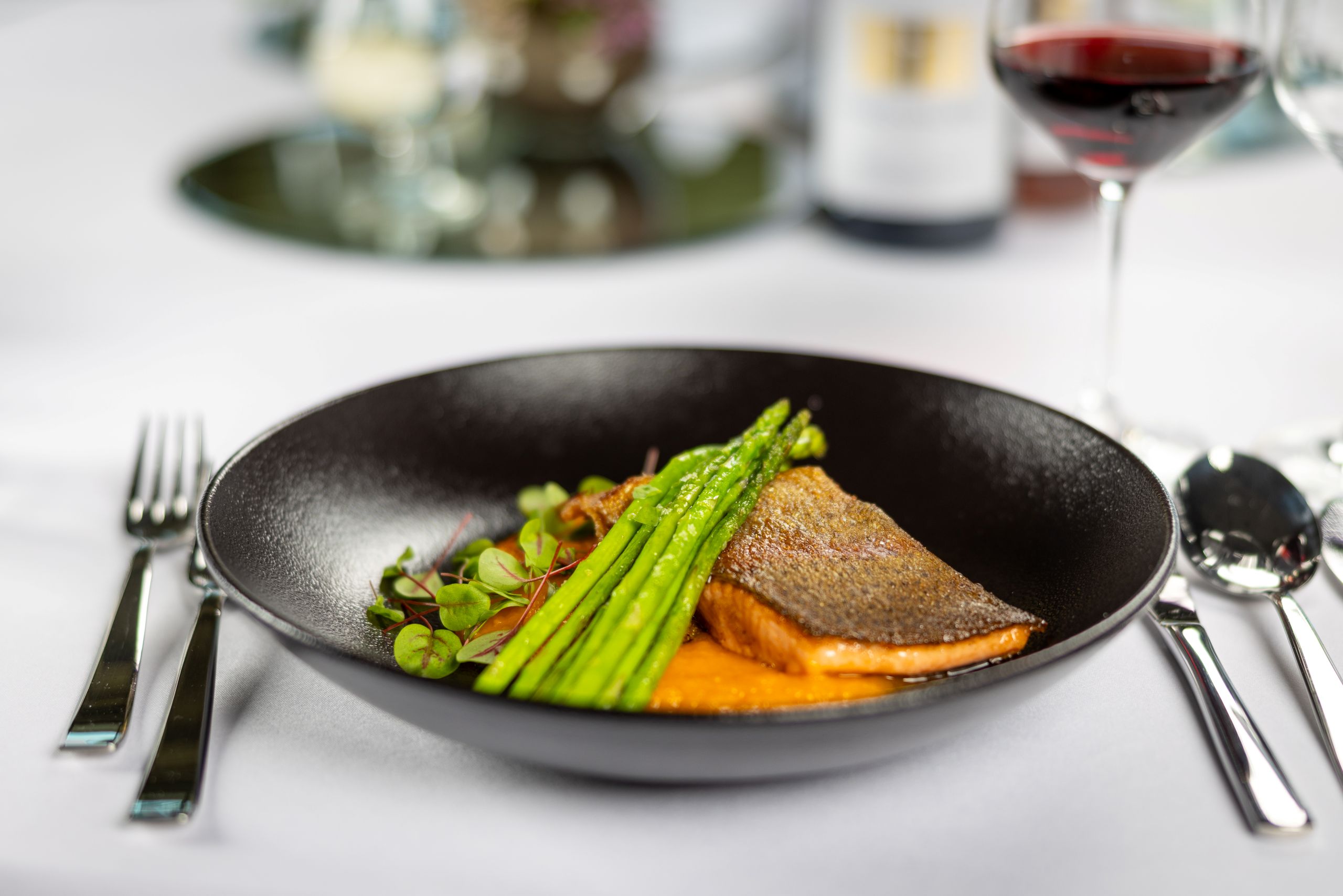 Gebratenes Stück Fisch mit krosser Haut auf orangfarbenem Püree, darüber liegt quer grüner Spargel und Kresse. Serviert in einer schwarzen flachen Schale auf einem gedeckten Tisch mit Besteck, Weingläsern und einer Flasche Wein im Hintergrund.