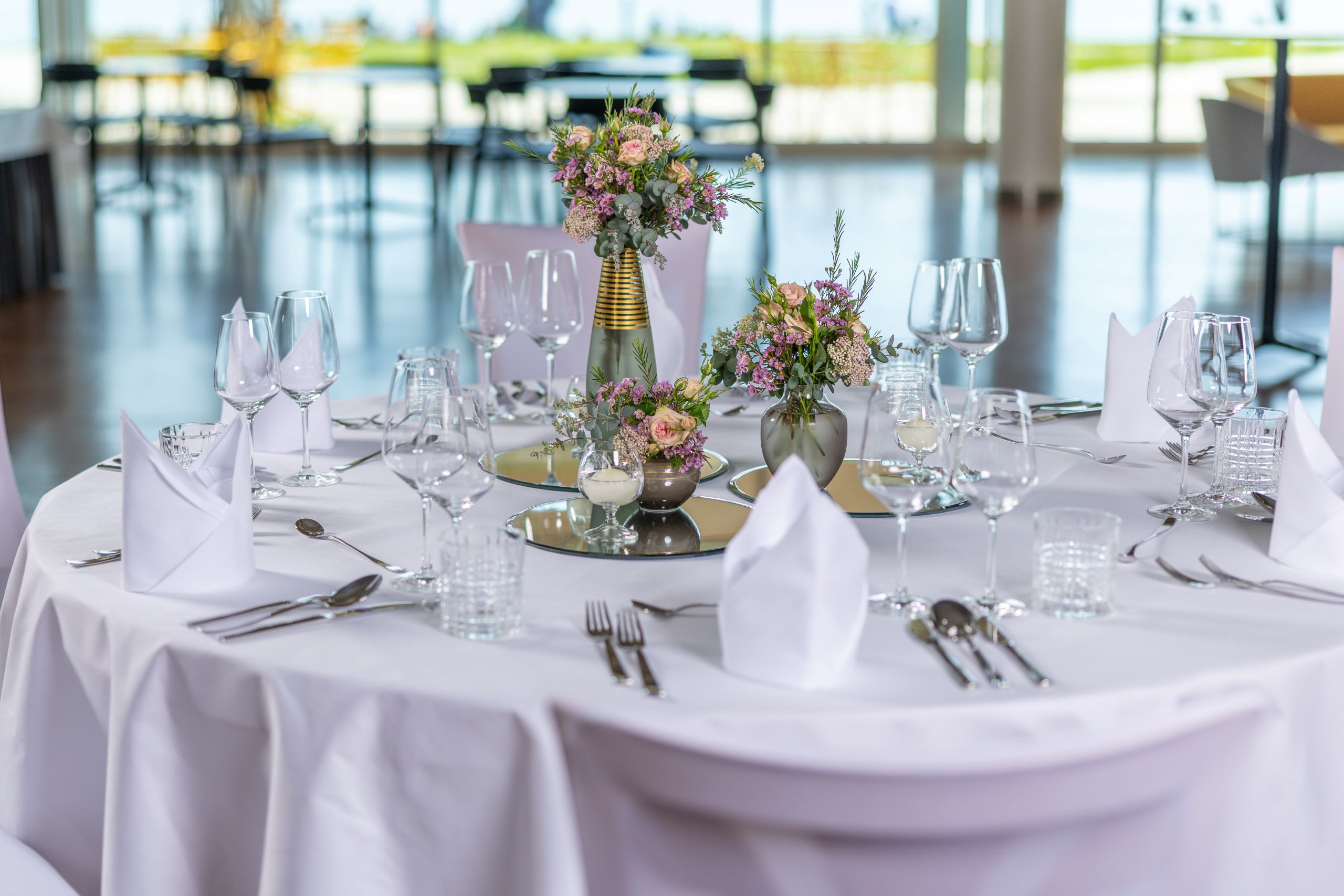 Feierlich eingedeckter Tisch mit weißer Tischdecke, Stoffservietten und Weingläsern, in der Mitte Blumendeko. Im Hintergrund sieht man verschommen ein Foyer mit dahinterligendem Außenbereich.